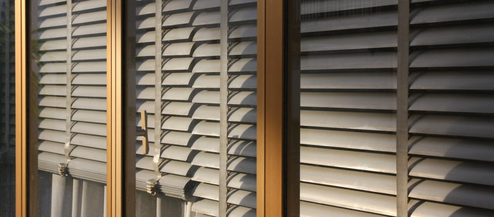 Finestre con persiane grigie e beige, viste attraverso montanti in legno e metallo, luce del sole.